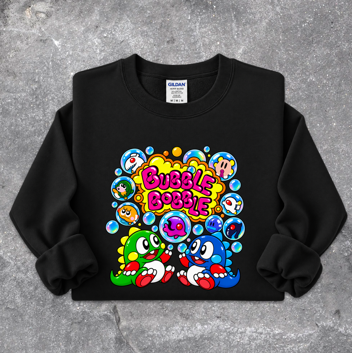 A black crewneck sweatshirt featuring a colorful retro cartoon graphic of two characters surrounded by bubbles with the text 'Bubble Bobble' in pink, laid flat on a gray concrete-textured background. The Gildan brand tag is visible inside the collar.