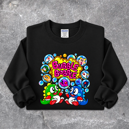 A black crewneck sweatshirt featuring a colorful retro cartoon graphic of two characters surrounded by bubbles with the text 'Bubble Bobble' in pink, laid flat on a gray concrete-textured background. The Gildan brand tag is visible inside the collar.