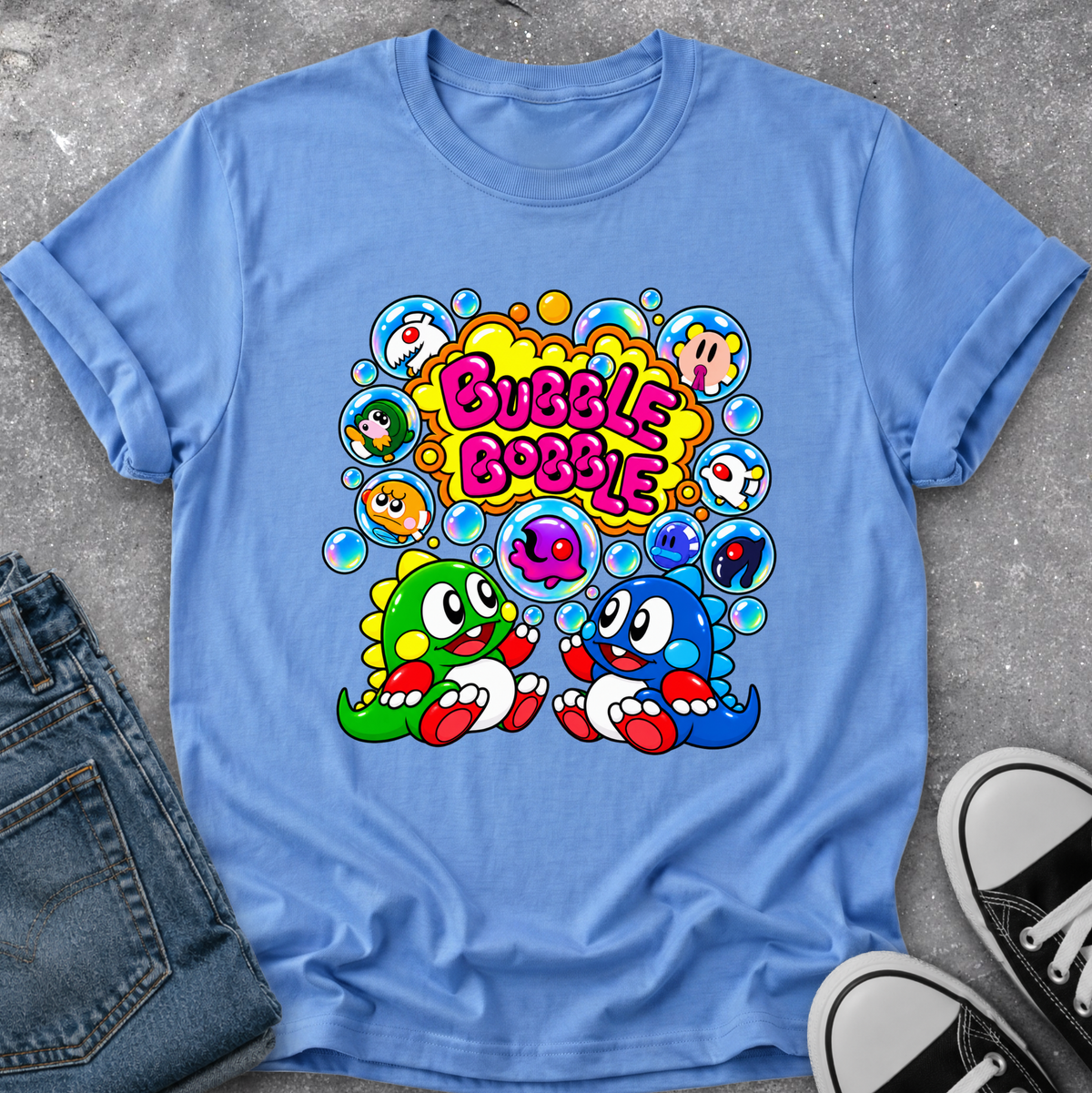 A light blue graphic t-shirt laid flat on a gray concrete surface, styled with a pair of blue jeans on the left and black-and-white sneakers on the right. The t-shirt features a colorful cartoon graphic of two dinosaur-like characters surrounded by bubbles with the text 'Bubble Bobble'.