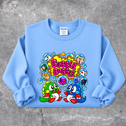 A light blue Gildan sweatshirt with a colorful cartoon graphic on the front, laid flat on a textured gray concrete background. The graphic features the text 'Bubble Bobble' in a yellow and pink bubbly font, surrounded by various colorful cartoon characters in circular bubbles, with two dinosaur-like characters at the bottom.