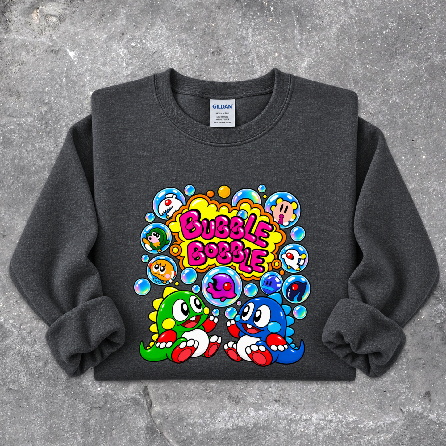 A dark gray crewneck sweatshirt featuring a colorful cartoon graphic of two cute dinosaur-like characters surrounded by bubbles with the text 'Bubble Bobble' in pink, laid flat on a textured gray concrete background. The neck label shows 'GILDAN'.
