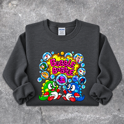 A dark gray crewneck sweatshirt featuring a colorful cartoon graphic of two cute dinosaur-like characters surrounded by bubbles with the text 'Bubble Bobble' in pink, laid flat on a textured gray concrete background. The neck label shows 'GILDAN'.
