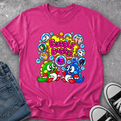 A bright pink short-sleeve t-shirt laid flat on a gray concrete surface, styled with blue jeans on the left and black-and-white sneakers on the bottom right. The shirt features a colorful retro graphic with the text 'Bubble Bobble' and two cartoon characters in green and blue surrounded by bubbles.