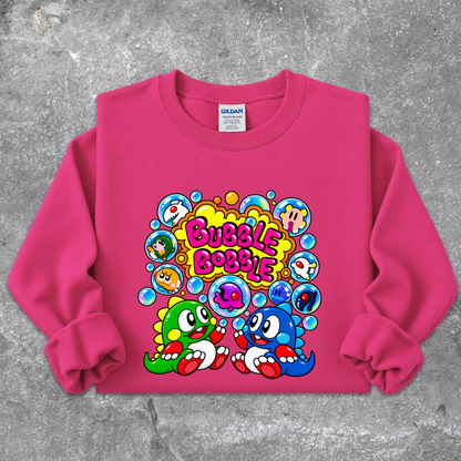 A bright pink crewneck sweatshirt featuring a colorful cartoon graphic of two green and blue characters with the text 'Bubble Bobble' in bubble letters, laid flat on a textured gray background.