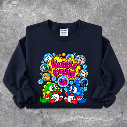 A navy blue crewneck sweatshirt with a colorful retro cartoon graphic on the front featuring the text 'Bubble Bobble' and two characters, laid flat on a textured gray concrete background. The neck label shows 'GILDAN'.
