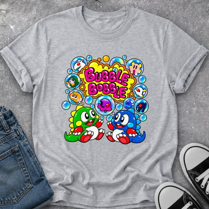 A heather gray short-sleeve t-shirt with a colorful retro graphic of two cartoon characters and the text 'Bubble Bobble'. The shirt is styled in a flat lay with blue jeans on the left and black-and-white sneakers on the right, set against a textured gray concrete background.