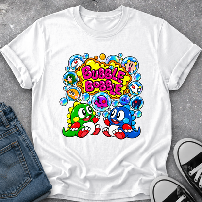 A white short-sleeve graphic T-shirt laid flat on a gray textured surface, styled with folded blue jeans on the left and black-and-white sneakers on the right. The shirt features a colorful retro-style print with the words 'Bubble Bobble' in pink/yellow bubble letters, surrounded by blue, green, and pink cartoon characters and bubbles.