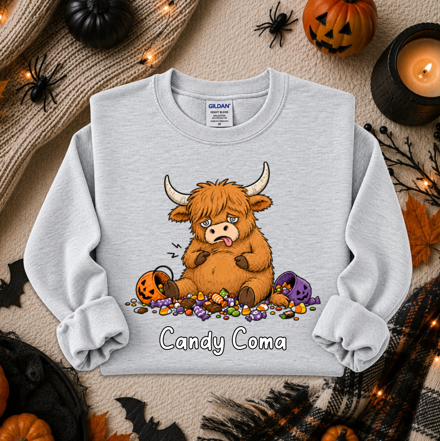 A light gray crewneck sweatshirt featuring a cartoon illustration of a fluffy brown Highland cow eating candy with the text 'Candy Coma' underneath. The sweatshirt is displayed in a flat-lay Halloween setting with pumpkins, a candle, and spider decorations.