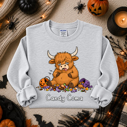 A light gray crewneck sweatshirt featuring a cartoon illustration of a fluffy brown Highland cow eating candy with the text 'Candy Coma' underneath. The sweatshirt is displayed in a flat-lay Halloween setting with pumpkins, a candle, and spider decorations.