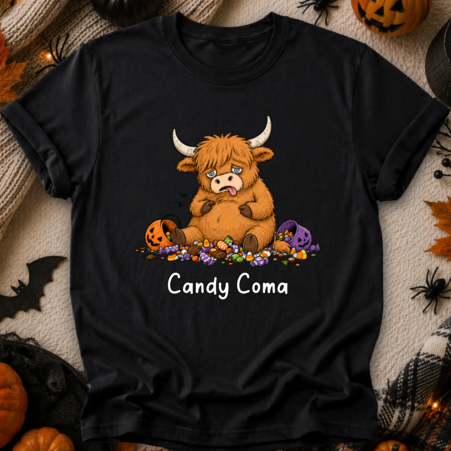 A black short-sleeve graphic T-shirt laid flat on a cozy beige fabric background with Halloween props. The shirt features a cartoon Highland cow surrounded by candy, with the text 'Candy Coma' below.