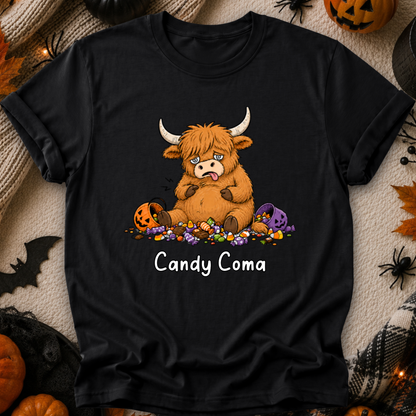 A black short-sleeve graphic T-shirt laid flat on a cozy beige fabric background with Halloween props. The shirt features a cartoon Highland cow surrounded by candy, with the text 'Candy Coma' below.
