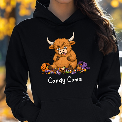 A person wearing a black hoodie with a cartoon graphic of a shaggy brown Highland cow sitting on candy, with pumpkins and a purple spider nearby, and the text 'Candy Coma' printed below. The background is a blurred outdoor scene with yellow autumn foliage.