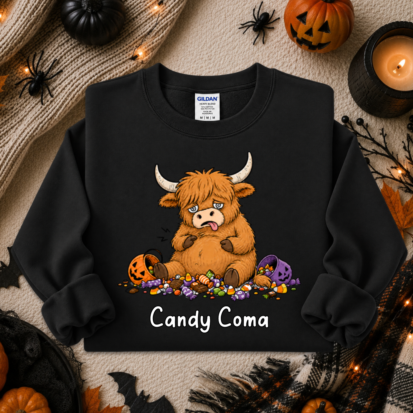 A black long-sleeve crewneck sweatshirt featuring a cartoon illustration of a fluffy orange Highland cow with white horns sitting on candy, with the text 'Candy Coma' printed below. The sweatshirt is displayed in a flat-lay Halloween setting with pumpkins, a candle, and spider decorations.