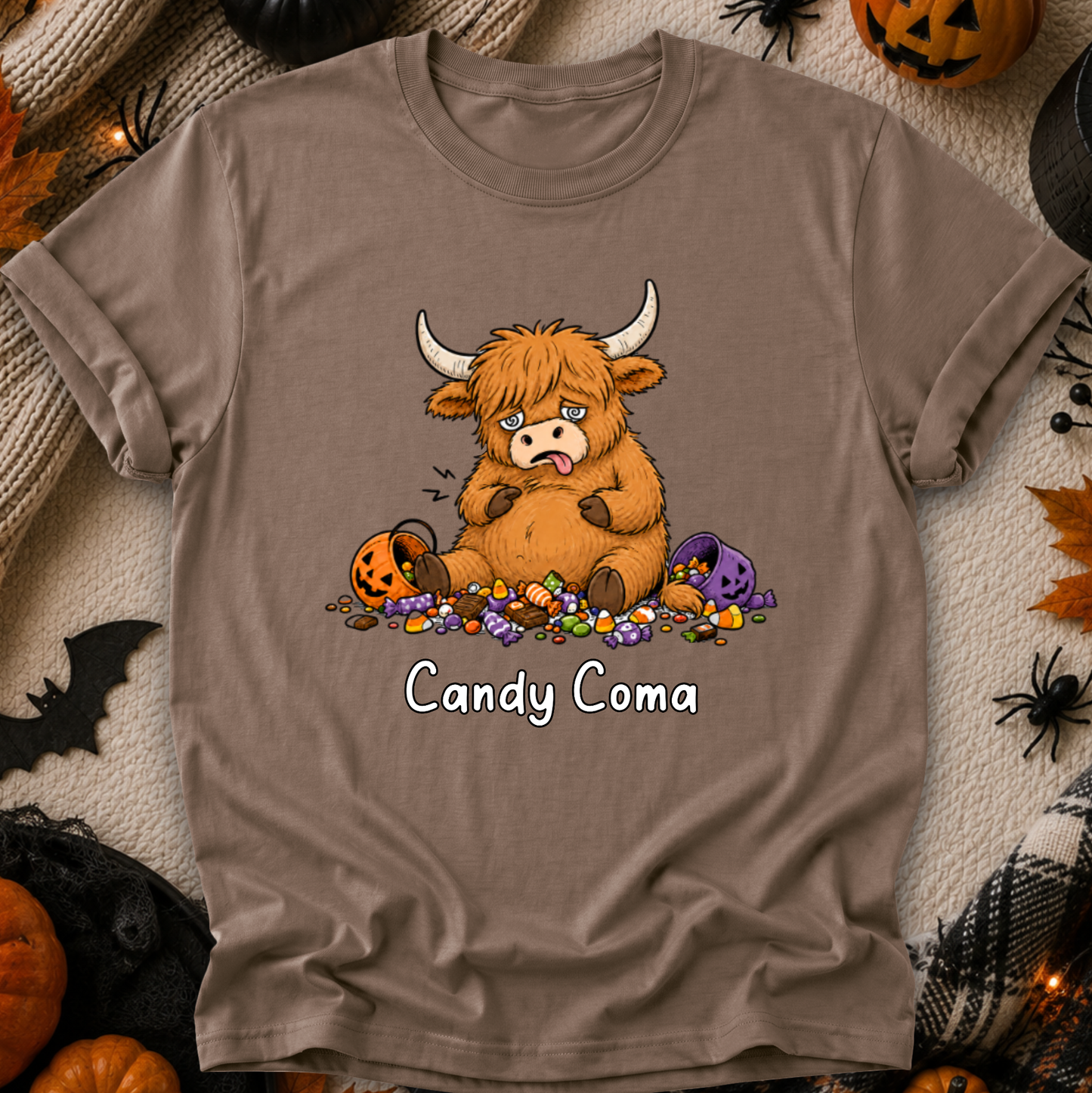 A taupe brown short-sleeve t-shirt with a cartoon graphic of a fluffy Highland cow surrounded by candy, with the text 'Candy Coma' printed below. The shirt is displayed in a flat-lay Halloween setting with pumpkins, spider decorations, and a cozy blanket background.