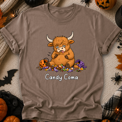 A taupe brown short-sleeve t-shirt with a cartoon graphic of a fluffy Highland cow surrounded by candy, with the text 'Candy Coma' printed below. The shirt is displayed in a flat-lay Halloween setting with pumpkins, spider decorations, and a cozy blanket background.