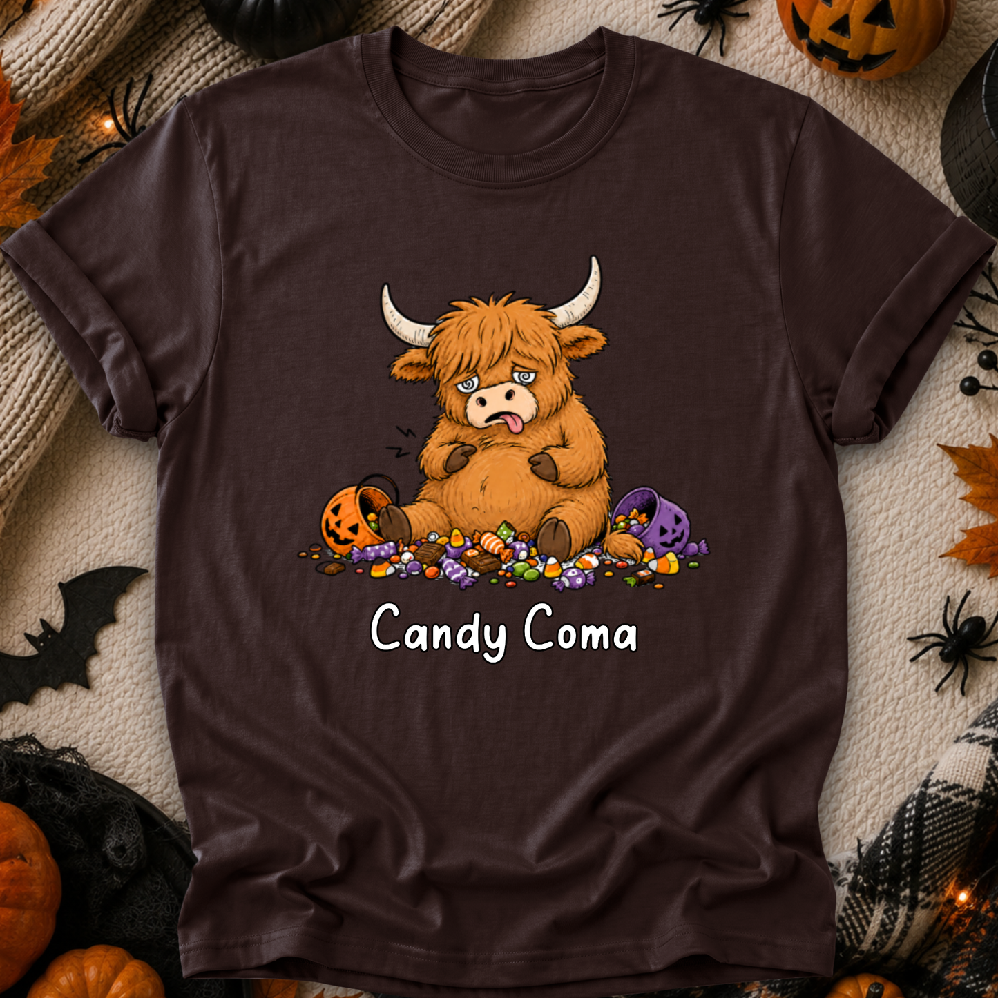 A dark brown short-sleeve t-shirt with a graphic of a cute cartoon Highland cow surrounded by candy and pumpkins, with the text 'Candy Coma' printed below. The shirt is displayed in a flat-lay Halloween setting with pumpkins, spider decorations, and a cozy blanket background.
