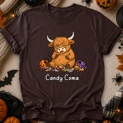 A dark brown short-sleeve t-shirt with a graphic of a cute cartoon Highland cow surrounded by candy and pumpkins, with the text 'Candy Coma' printed below. The shirt is displayed in a flat-lay Halloween setting with pumpkins, spider decorations, and a cozy blanket background.