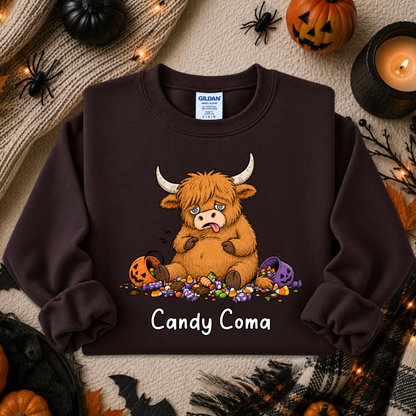 A dark brown crewneck sweatshirt featuring a cartoon graphic of a fluffy orange Highland cow sitting on candy with the text 'Candy Coma' underneath. The sweatshirt is displayed in a flat-lay Halloween setting with pumpkins, a candle, and spider decorations.