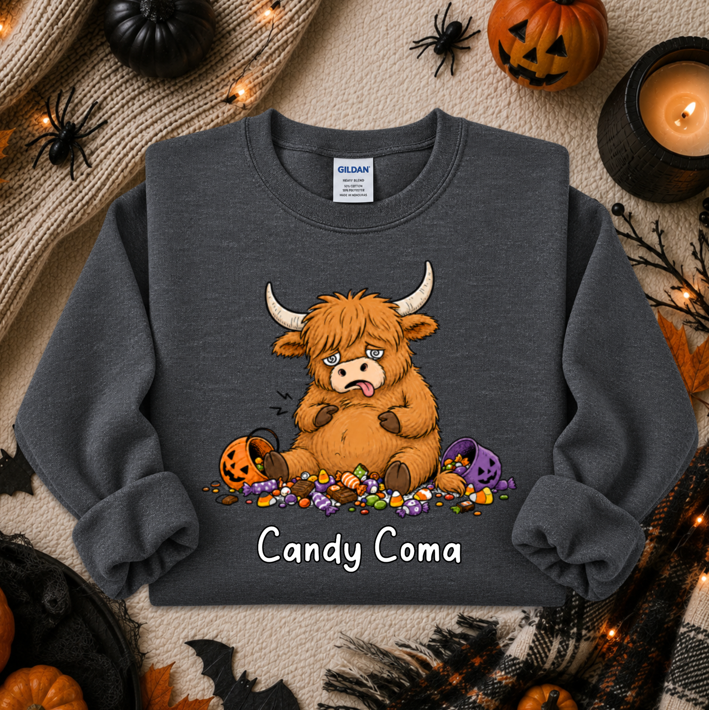 A dark gray crewneck sweatshirt featuring a cartoon illustration of a fluffy orange Highland cow eating candy, with the text 'Candy Coma' printed below. The sweatshirt is displayed in a flat-lay Halloween setting with pumpkins, a candle, and spider decorations.