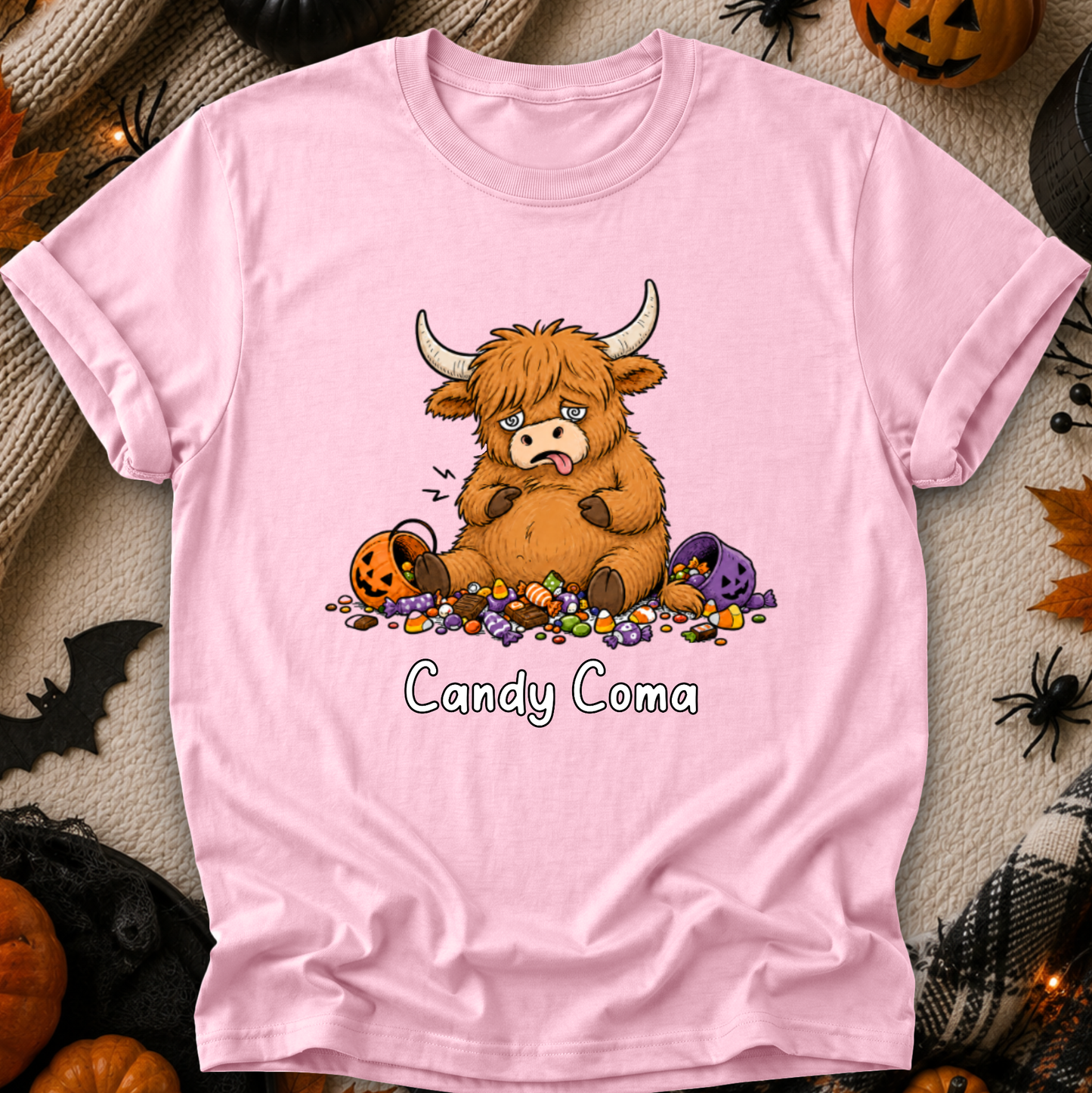 A light pink short-sleeve t-shirt with a cartoon graphic of a sad, tired Highland cow surrounded by candy, with the text 'Candy Coma' printed below. The shirt is displayed flat on a Halloween-themed background with pumpkins, spiders, and a bat.