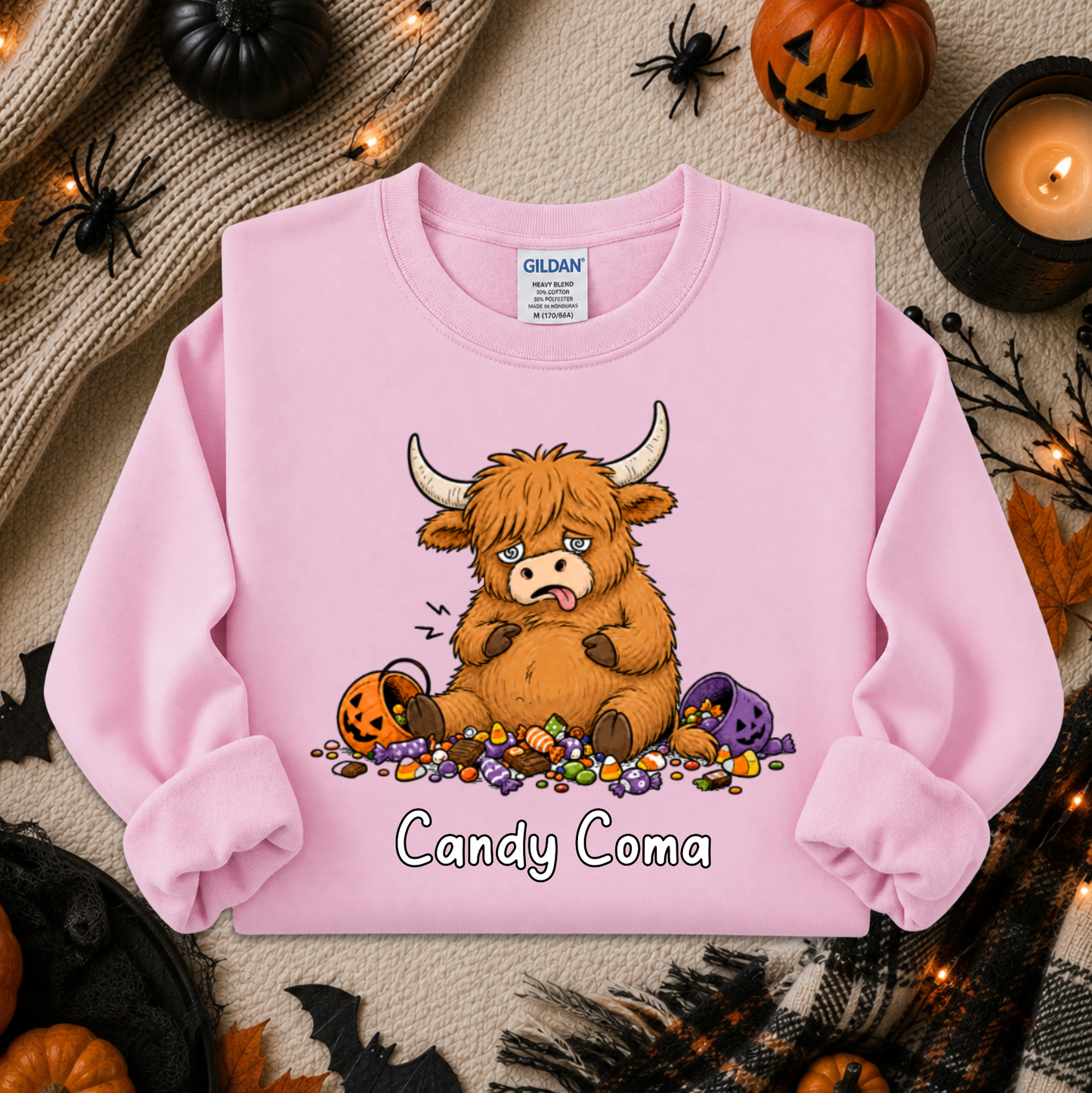 A pink long-sleeve sweatshirt laid flat on a cozy autumn-themed background. The sweatshirt features a cartoon illustration of a fluffy brown Highland cow eating candy, with the text 'Candy Coma' printed below.