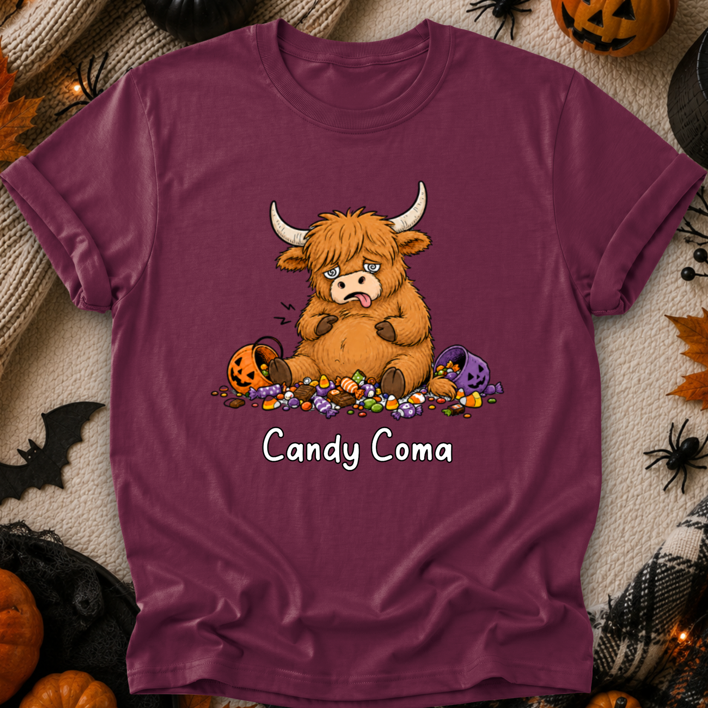 A mauve-purple short-sleeve t-shirt with a cartoon Highland cow graphic surrounded by candy and the text 'Candy Coma', displayed in a flat-lay Halloween setting with pumpkins, spiders, and a black spiderweb.