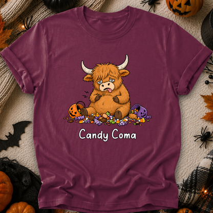 A mauve-purple short-sleeve t-shirt with a cartoon Highland cow graphic surrounded by candy and the text 'Candy Coma', displayed in a flat-lay Halloween setting with pumpkins, spiders, and a black spiderweb.