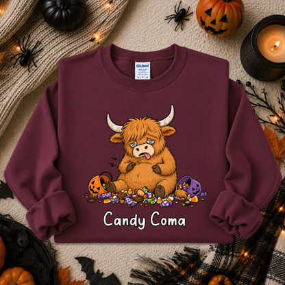 A maroon crewneck sweatshirt featuring a cartoon illustration of a fluffy brown Highland cow surrounded by candy and pumpkins, with the text 'Candy Coma' printed below. The sweatshirt is displayed in a flat-lay Halloween setting with a knit sweater, candles, and jack-o'-lanterns.
