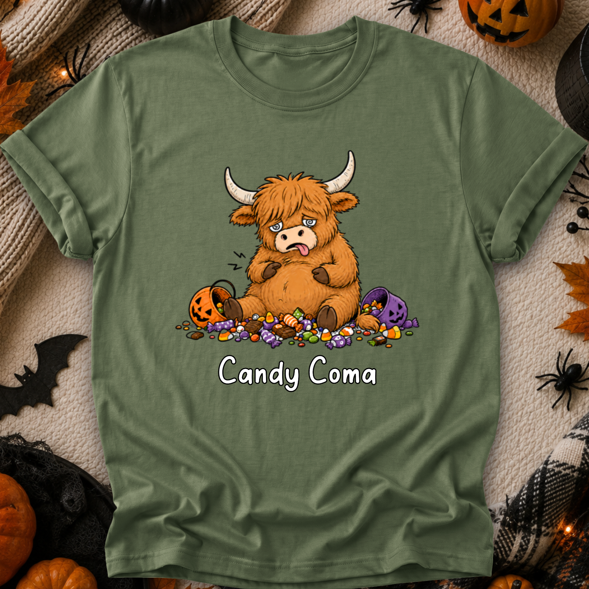 An olive green short-sleeve t-shirt with a cartoon graphic of a tired Highland cow surrounded by candy and a pumpkin, with the text 'Candy Coma' printed below. The shirt is displayed flat on a cozy, decorated Halloween-themed background.