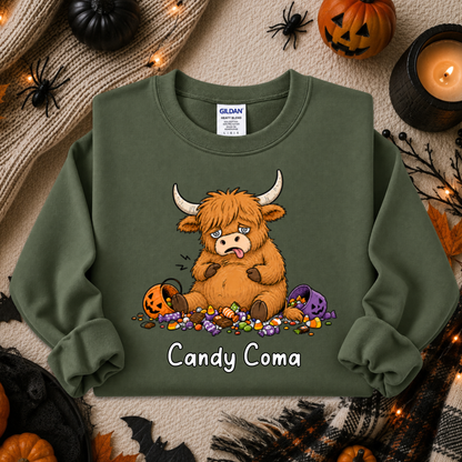 An olive green crewneck sweatshirt featuring a cartoon graphic of a fluffy brown Highland cow eating candy with the text 'Candy Coma' underneath. The sweatshirt is displayed in a flat-lay Halloween setting with pumpkins, a candle, and spider decorations.