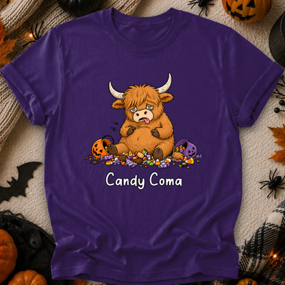 A purple short-sleeve t-shirt with a cartoon graphic of a sad Highland cow surrounded by candy and jack-o'-lanterns, with the text 'Candy Coma' printed below. The shirt is displayed flat on a cozy, Halloween-themed background with pumpkins, a knitted blanket, and black spider decorations.