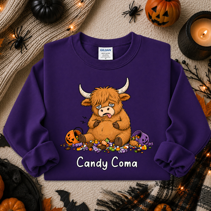 A purple long-sleeve crewneck sweatshirt featuring a cartoon Highland cow surrounded by candy and pumpkins, with the text 'Candy Coma' printed below. The sweatshirt is displayed in a flat-lay Halloween setting with a knit sweater, candles, and jack-o'-lanterns.