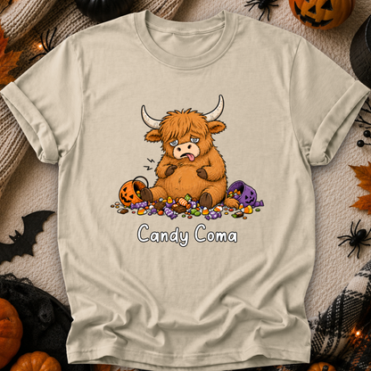 A beige graphic t-shirt featuring a cartoon Highland cow eating candy, surrounded by Halloween props like pumpkins, spiders, and a black plaid blanket, with the text 'Candy Coma' printed below the graphic.