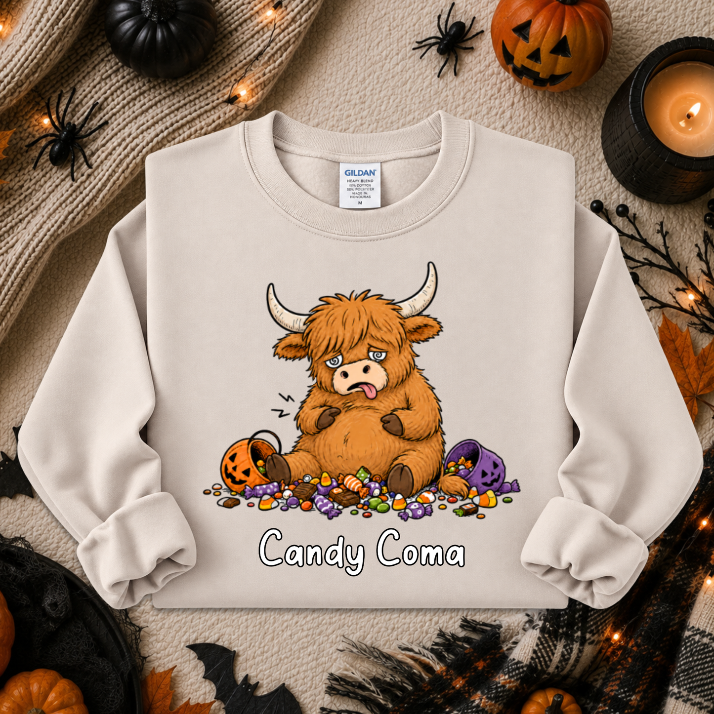 A cream-colored crewneck sweatshirt featuring a cartoon illustration of a fluffy brown Highland cow with its tongue out, surrounded by candy corn, purple pumpkins, and the text 'Candy Coma'. The sweatshirt is displayed in a flat-lay Halloween setting with pumpkins, a candle, and a knit blanket.