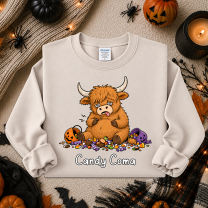 A cream-colored crewneck sweatshirt featuring a cartoon illustration of a fluffy brown Highland cow with its tongue out, surrounded by candy corn, purple pumpkins, and the text 'Candy Coma'. The sweatshirt is displayed in a flat-lay Halloween setting with pumpkins, a candle, and a knit blanket.