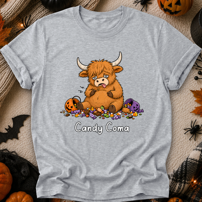 A light heather gray short-sleeve t-shirt with a cartoon graphic of a fluffy brown Highland cow surrounded by candy, with the text 'Candy Coma' underneath. The shirt is displayed in a flat-lay Halloween setting with pumpkins, a spider, and a black spiderweb.