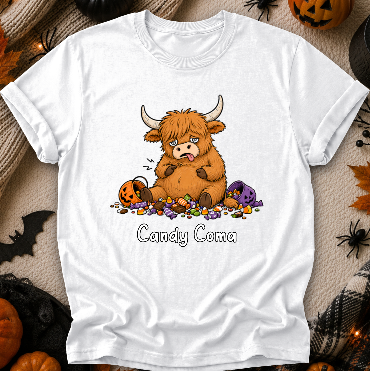 A white short-sleeve crewneck t-shirt featuring a cartoon graphic of a fluffy brown Highland cow sitting in a pile of colorful candy, with the text 'Candy Coma' printed below. The shirt is displayed in a flat-lay Halloween setting with pumpkins, spider decorations, and a black bat silhouette.