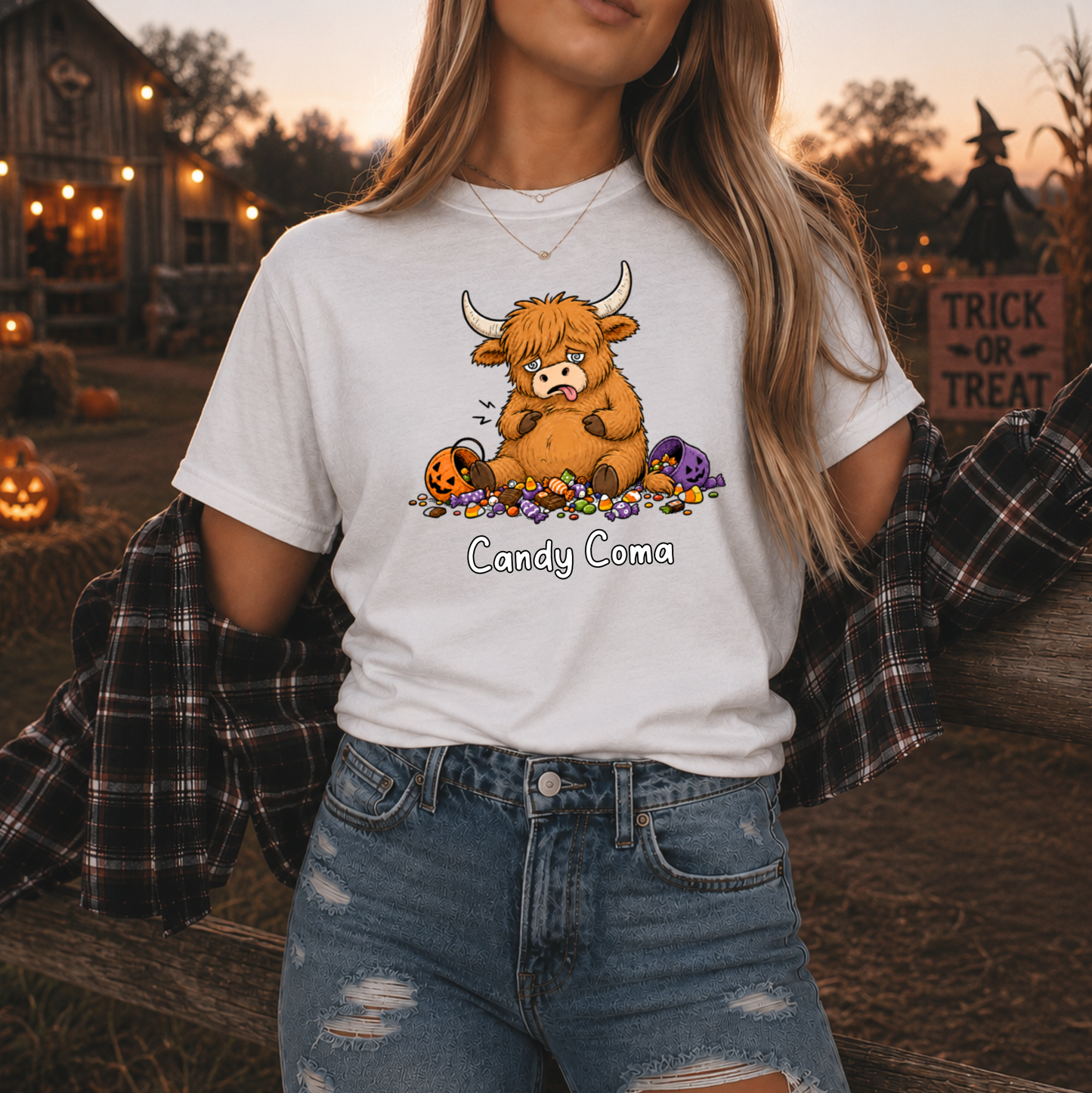A woman wearing a white graphic t-shirt with a cartoon Highland cow surrounded by candy corn and purple pumpkins, with the text 'Candy coma' printed below. She is wearing a black plaid flannel shirt off her shoulders and distressed blue jeans, set against a warm, festive autumn background with pumpkins and a rustic barn.