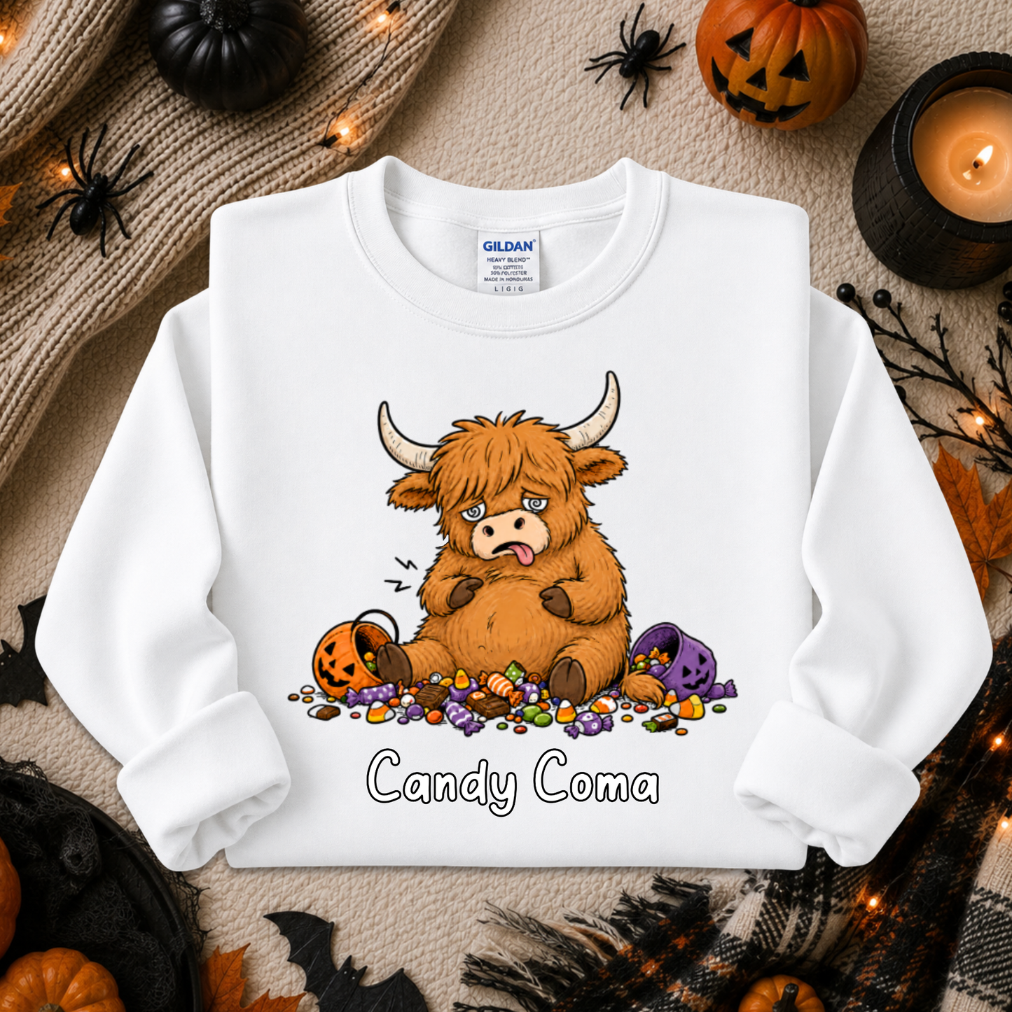 A white long-sleeve crewneck sweatshirt with a cartoon graphic of a tired, comatose Highland cow surrounded by candy, with the text 'Candy Coma' printed below. The sweatshirt is displayed in a flat-lay arrangement with Halloween props including pumpkins, a candle, and a knit sweater.