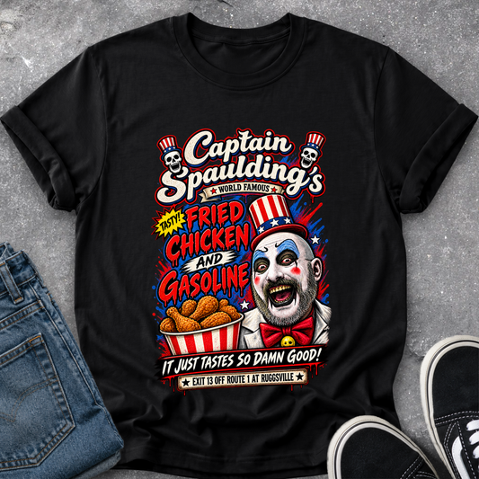 A black graphic t-shirt featuring a colorful retro clown design labeled 'Captain Spaulding's' with fried chicken and gasoline text, styled flat on a gray concrete surface with blue jeans and black sneakers partially visible.