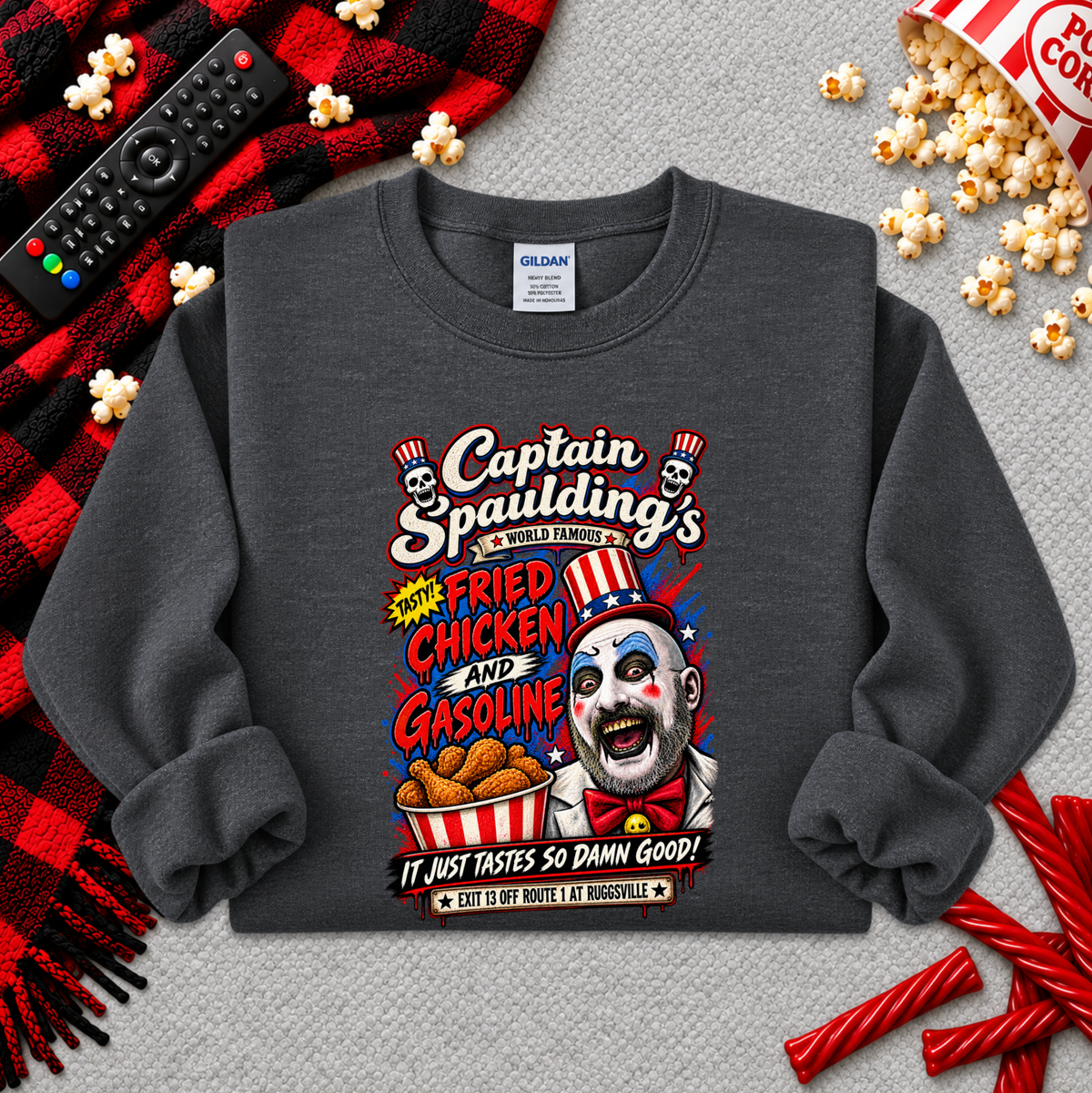 A dark gray crewneck sweatshirt featuring a colorful graphic print of a clown with the text 'Captain Spaulding's Fried Chicken and Gasoline'. The sweatshirt is styled in a flat-lay arrangement with a red and black plaid blanket, a remote control, popcorn, and candy on a gray textured background.
