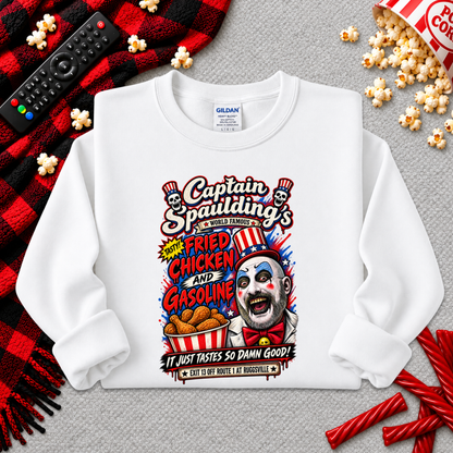 A white long-sleeve crewneck sweatshirt with a colorful graphic print of 'Captain Spaulding's Fried Chicken and Gasoline' clown character, styled in a flat lay with popcorn, candy, a remote, and a red plaid blanket on a gray textured background.