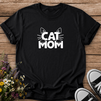 A black short-sleeve t-shirt with a white graphic reading 'CAT MOM' and cat ears/paws, laid flat on a wooden surface with flowers and black-and-white sneakers as props.