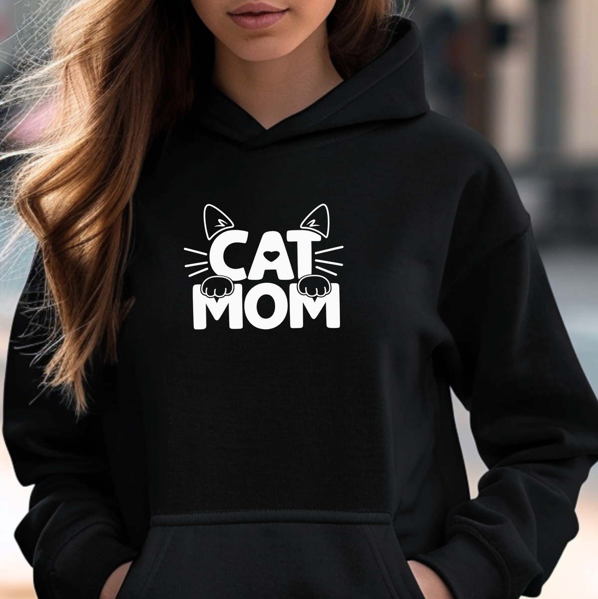 A woman wearing a black pullover hoodie with a white graphic print that reads 'CAT MOM' with cat ears and paw prints, shown in a close-up lifestyle shot.