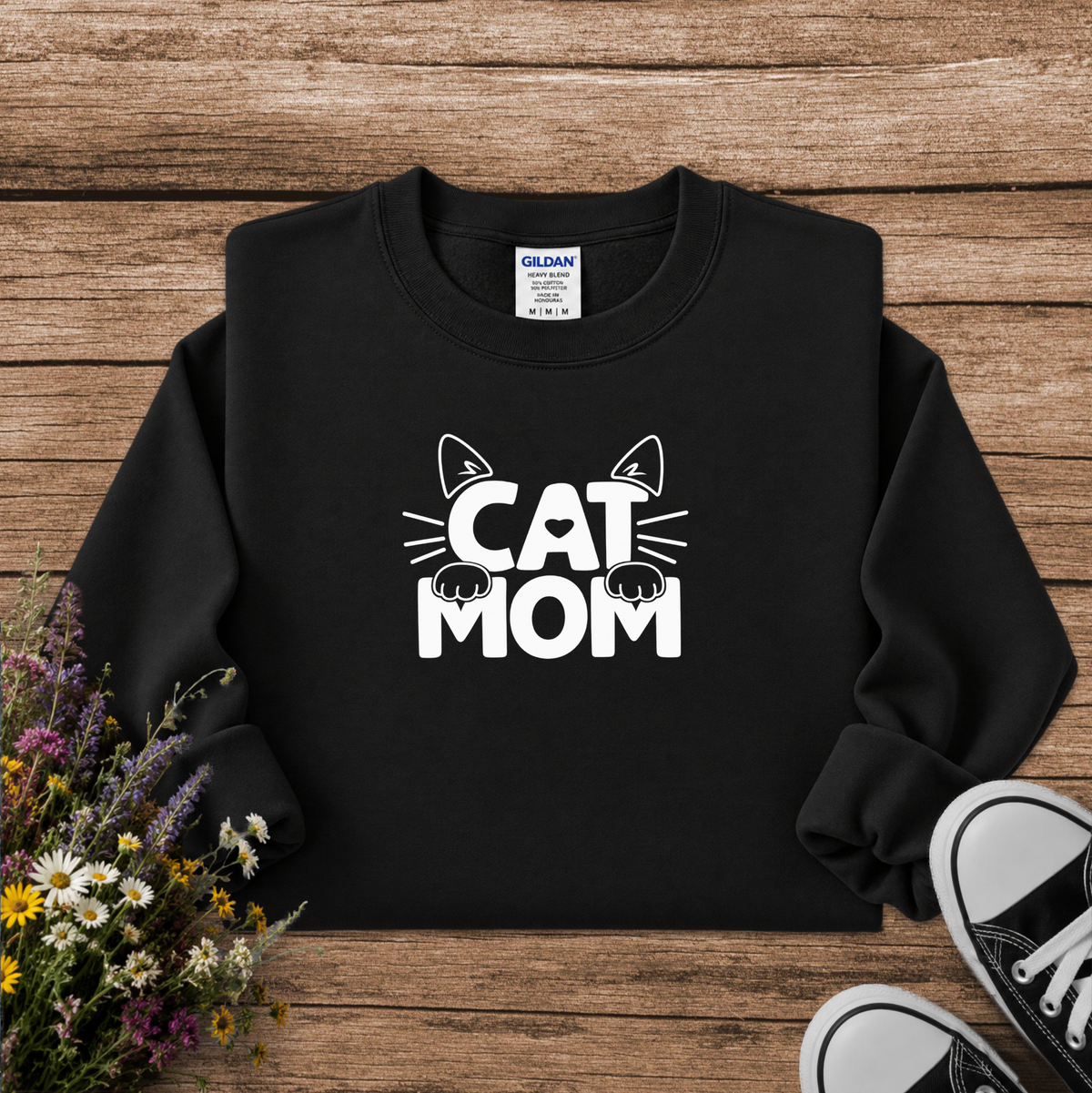 A black crewneck sweatshirt with a white graphic reading 'CAT MOM' and cat ears/paws, laid flat on a rustic wood background with flowers and black sneakers as props. A Gildan neck label is visible.