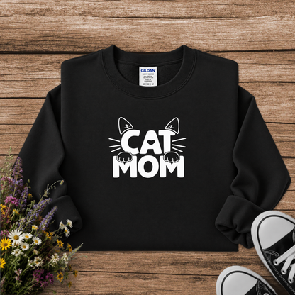 A black crewneck sweatshirt with a white graphic reading 'CAT MOM' and cat ears/paws, laid flat on a rustic wood background with flowers and black sneakers as props. A Gildan neck label is visible.