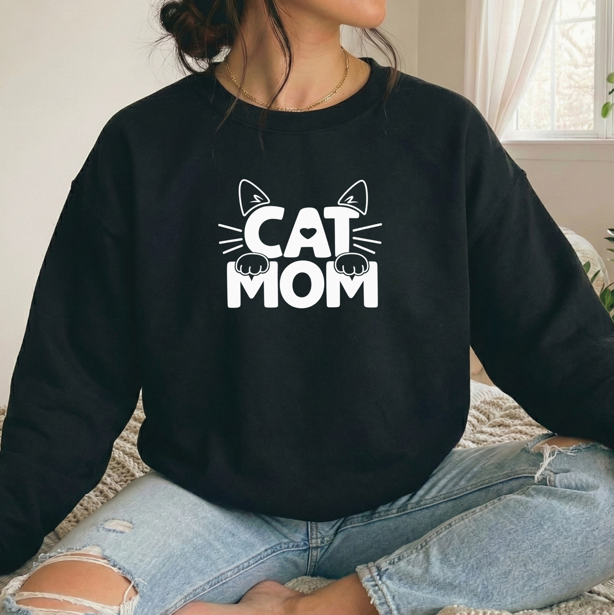 A person wearing a black crewneck sweatshirt with a white 'CAT MOM' graphic featuring cat ears and paws, paired with light blue ripped jeans in a bright indoor setting.
