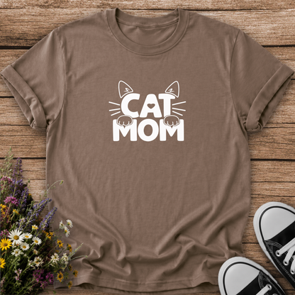 A brown short-sleeve t-shirt with a white graphic reading 'CAT MOM' and cat ears/paws, laid flat on a wooden surface with flowers and black-and-white sneakers as props.