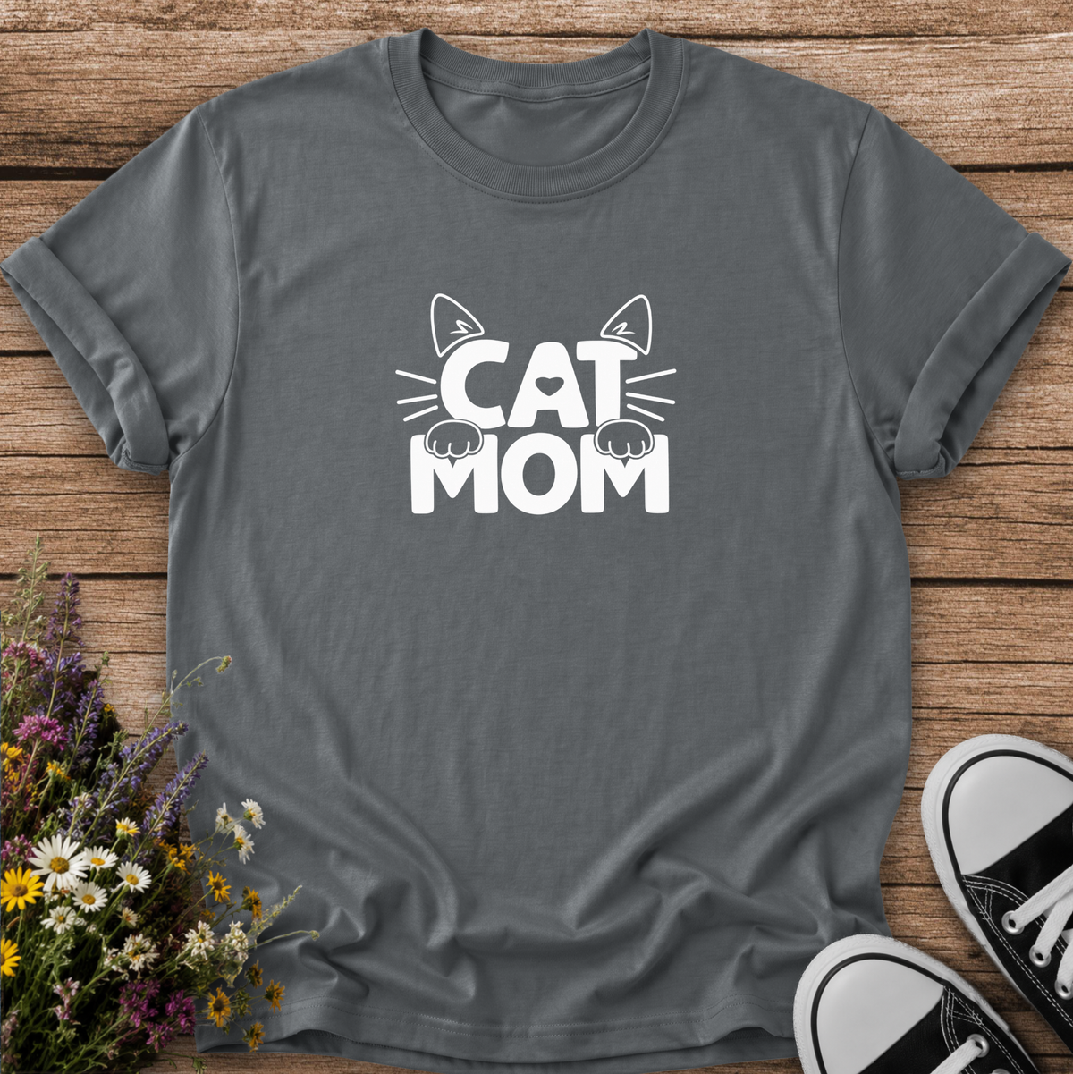 A dark gray crewneck t-shirt with a white graphic reading 'CAT MOM' featuring cat ears, whiskers, and paws, laid flat on a rustic wooden background with wildflowers and black-and-white sneakers.