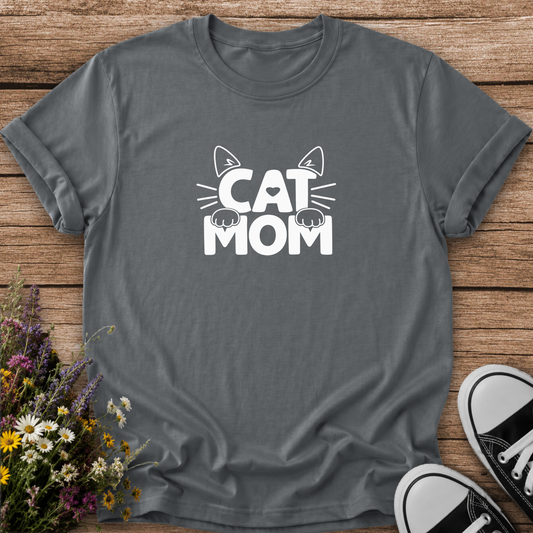A dark gray crewneck t-shirt with a white graphic reading 'CAT MOM' featuring cat ears, whiskers, and paws, laid flat on a rustic wooden background with wildflowers and black-and-white sneakers.